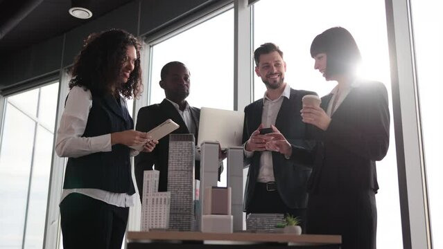 Diverse group of multiracial business people in the conference room with panoramic city view cooperating standing near table with 3d skyscraper building maquette. Teamwork concept.