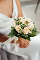 Bride woman holds a wedding bouquet in her hands on the background of white dress. Wedding ceremony. Beautiful beige roses flower and green leaves. Tender sensual close up