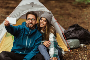Glad millennial caucasian husband and wife resting in cold forest, enjoy adventure, sit in tent, taking selfie