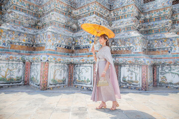 Young woman wearing Thai dress with accessories stands holding antique umbrella and bag at Wat Arun Ratchawararam It is a popular destination for tourists around the world. Bangkok, Thailand