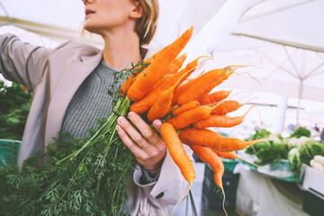 Woman with organic carrots buying vegetables and greens in local market. People doing shopping of...