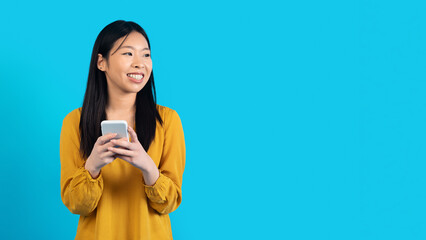 Cheerful young asian woman using phone on blue, copy space