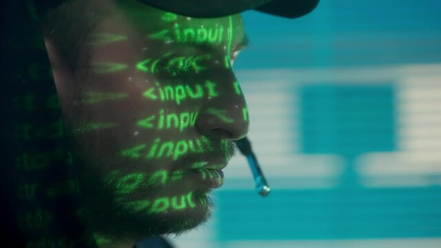 Hacker with headset working on a computer, a line of program code is reflected on his face. A green neon source code is projected onto a close up profile of a man's face against a blue background.