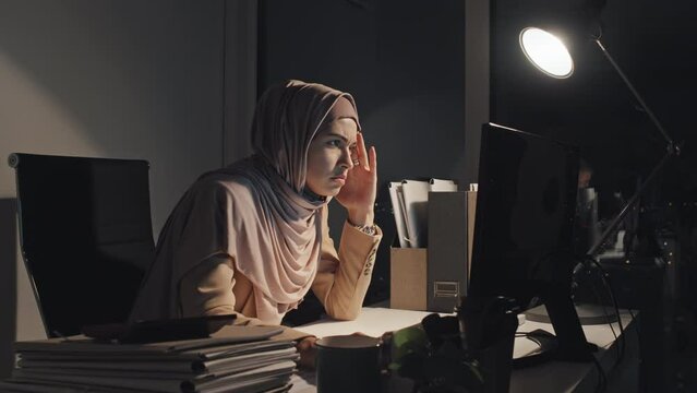 Medium shot of young Muslim woman in hijab sitting at desk under bright lamp at night in office, typing on keyboard, reading documents on computer, then looking at wristwatch, sighing and shaking head