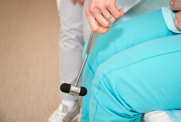 Pretty female orthopedist with patient in her office. Young doctor neurologist examining her patient with hammer. Neurologist checks the reflexes of a middle age female patient. Close up view.