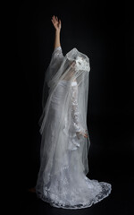Full length portrait of beautiful woman wearing  white bridal fantasy  gown with butterfly mask and ethereal veil.  Dancing with gestural arm poses, isolated on black studio background.