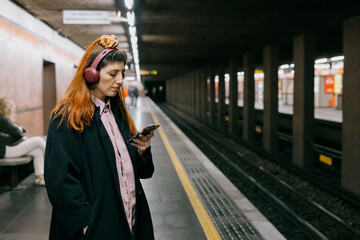 Bored young woman waits for subway listening music in headphones from smart phone