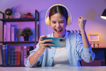 Excited Young Asian woman wearing headset and playing online game on smartphone at night modern house. © sitthiphong