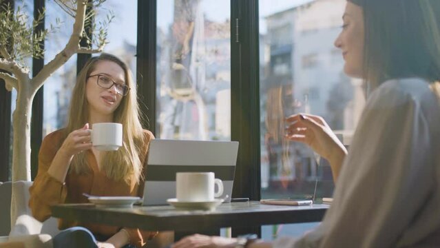 Two young business women in a cafe having one on one meeting. Friends after work talking gossiping and having coffee at a window table with reflections.