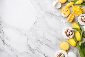 Set of easter sweets with yellow spring flowers and eggs over marble background. Top view, copy space, frame composition. Meringues in shape of nest, macaroon chicks, daffodils and tulips