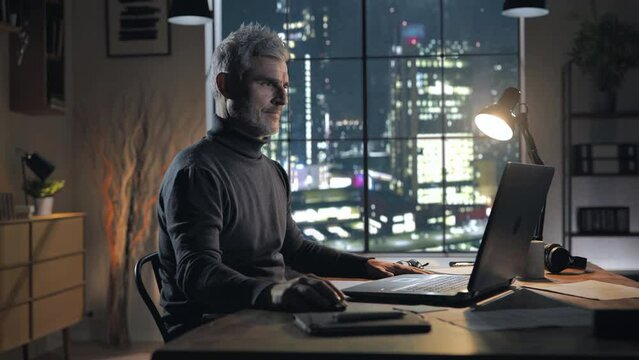 Side view of focused mature male working late at the computer from home office at night,handsome middle age man sits at the desk works at the laptop in modern studio window city view in the background