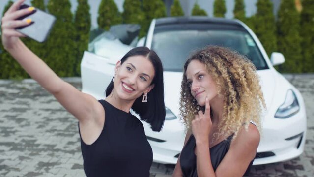 Two beautiiful girls in black outfut smiling and posing for a selfy photo in front of fancy sports car..