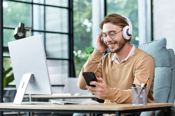 Smiling young male programmer, freelancer sits in the office at the table in headphones and holds...