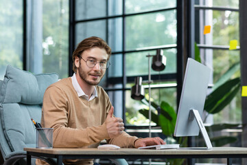 Portrait of a young man in glasses working at a computer in a modern office. He looks at the camera, smiles, shows a successful gesture with a super finger.