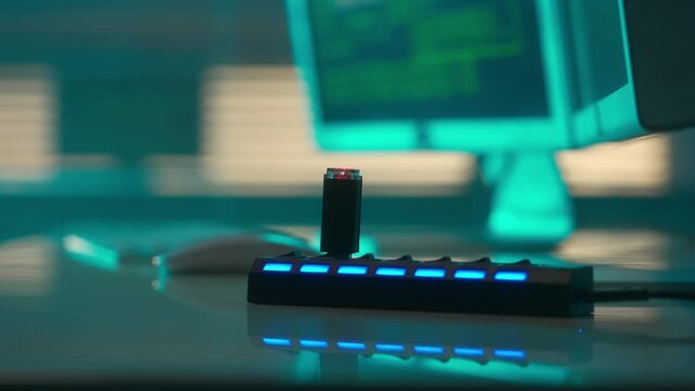 A black flash drive with a red flashing light is inserted into the USB port. USB flash drive in a card reader against the background of computer displays with green lines of codes moving on screens.