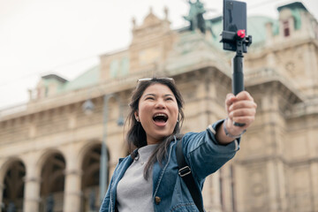 Young casual woman using smartphone to record journey with smile. Happy and excited asian girl traveler wear stylish cloth with hand gesture waving to internet audience. Influencer vlogging concept