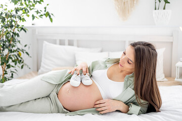 Obraz na płótnie Canvas A pregnant woman with a big open belly holds socks and dreams of having a baby. The expectant mother is waiting and preparing for the birth of a child in a bright bedroom
