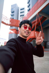 young asian man wearing cool red glasses taking point of view selfie waving and smiling