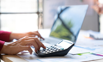 Asian businesswoman recording data on laptop Analyze charts, graphs and calculate financial reports with calculator in financial advisory office and accounting concept.