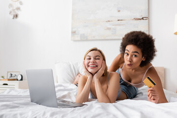 african american woman holding credit card and grimacing near cheerful blonde friend and laptop on bed.