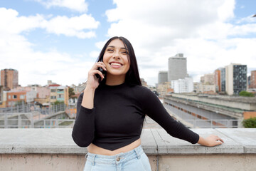 Fototapeta premium Panoramic view of a beautiful smiling woman talking on the phone on a terrace in Bogota with the cityscape in the background, Colombia.