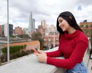 Fototapeta premium smiling woman using smartphone. Girl with black hair and red jacket smiles using smartphone.