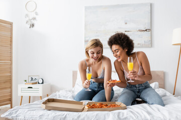 Obraz na płótnie Canvas happy interracial women holding champagne glasses with cocktails while looking at delicious pizza on bed at home.