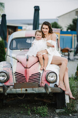 Fototapeta na wymiar A beautiful mother with a daughter in a white dress posing near red retro car. Mom with child enjoying day. Summer family photo. Spring fashion model concept. Vintage and retro style. Luxury travel.