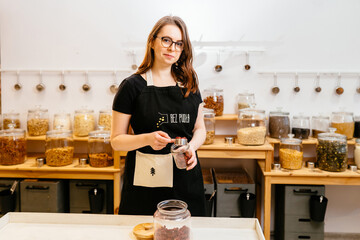 Confident female owner holding spice jar by rack at zero waste store. Eco-friendly shopping at local small businesses New trend alternative buying.