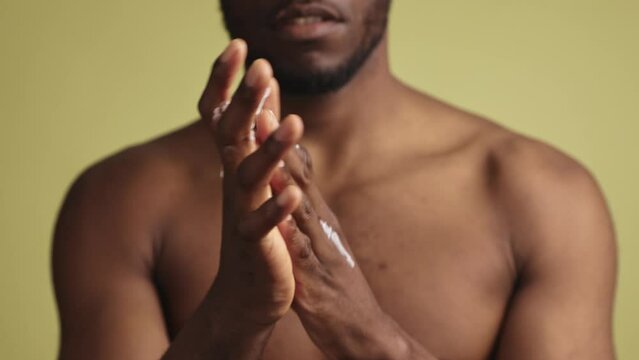 Close-up of handsome sensitive young black man applying hand cream looking at hands on khaki green background. The concept of male grooming and beauty.