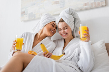 young and happy multiethnic women in white bathrobes and towels holding fruits and fresh orange juice in bedroom.