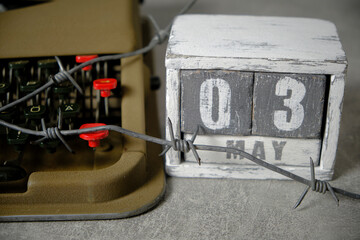 Typewriter is wrapped with barbed wire with wooden calendar next with date May 3.Concept Press...