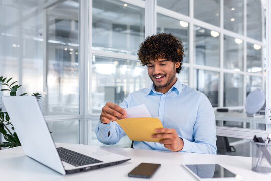 Young successful businessman inside office working with laptop, man received good news notification mail, hispanic man reading news and smiling at workplace, happy with achievement result.