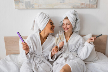 joyful multiethnic women in white terry robes and towels holding nail files and clinking champagne glasses on bed.