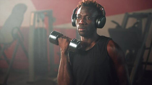muscular black man with headphones is training alone in fitness gym, lifting dumbbells
