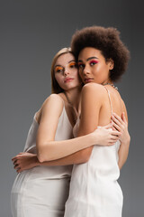 multiethnic blonde and brunette models in white camisoles and bright visage embracing isolated on grey.