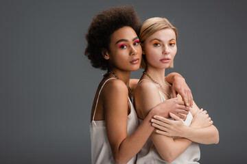 multiethnic blonde and brunette models with colorful visage looking at camera isolated on grey.