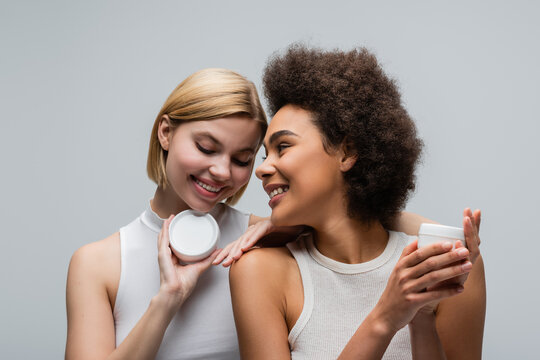 young and pretty multiethnic women in white tops posing with jars of body cream isolated on grey.