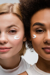 close up view of young interracial women with face cream smears isolated on grey.