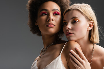 blonde and brunette interracial women with bright visage and silver necklaces posing isolated on grey.