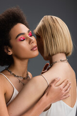 sensual african american model with glossy eye shadows and closed eyes embracing blonde woman isolated on grey.