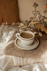 Hot mug of cappuccino on wooden tray on the bed, breakfast, bouquet of dried flowers. Spring scenery. Cozy house. Beige natural colors.