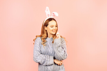 Obraz na płótnie Canvas Beautiful girl in a blue sweater on a pink background. Woman with bunny ears, posing for easter, girl smiling, space for text.