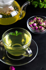 cup of tea with flowers. Rose buds tea in a glass teapot on the black table. Teapot with tea made from tea rose petals with green mint.