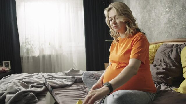 Lifestyle video of a pregnant woman at home watching TV shows on a tablet and eating candy. Cheerful smiling girl with pregnant belly eating sweets on bed.