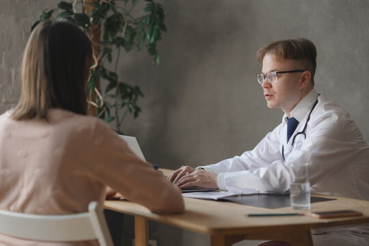 Man Doctor Communicates With Female Patient In Office And Works Laptop In Clinic. Male Therapist At Reception Of Case Prescribes Treatment With Medication. Virologist, Immunologist, Endocrinologist