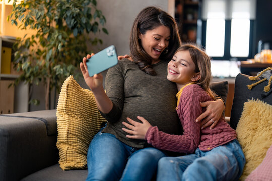 Cheerful mother and smiling daughter using mobile phone while sitting and resting on sofa at home, taking selfies or having a video call with family or friends.