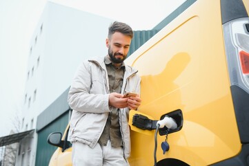 Fototapeta na wymiar bearded caucasian man standing near an electric car that is charging and making time adjustments on a smartphone.