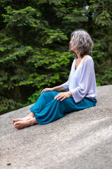 Femme qui médite, assise sur un gros rocher de granit, vêtue d'un pantalon bleu et d'un petit pull violet 