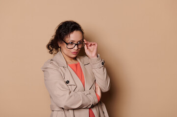 Isolated portrait on beige background of a multi-ethnic middle-aged curious brunette lady, holding stylish spectacles, looking at empty space for advertising text. Health. Fashion. Eyesight problems